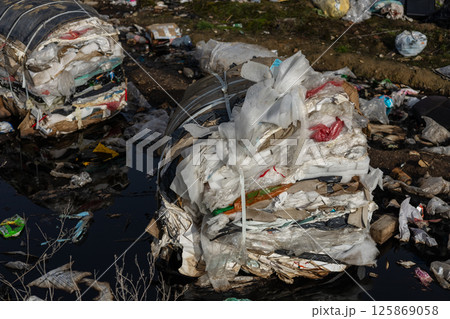 Large piles of compressed waste materials are seen near a polluted water source in an urban setting, highlighting environmental concerns Large piles of compressed waste materials are seen near a polluted water source in an urban setting, highlighting environmental concerns 125869058