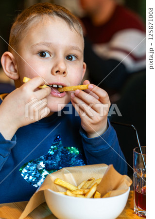 Happy boy with french fry in mouth, eating fries from bowl, indoor casual restaurant background, concept of joyful childhood meal 125871008