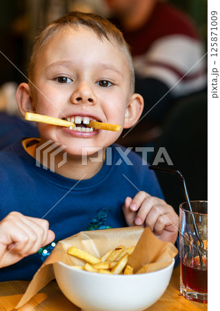 Happy boy with french fry in mouth, eating fries from bowl, indoor casual restaurant background, concept of joyful childhood meal 125871009