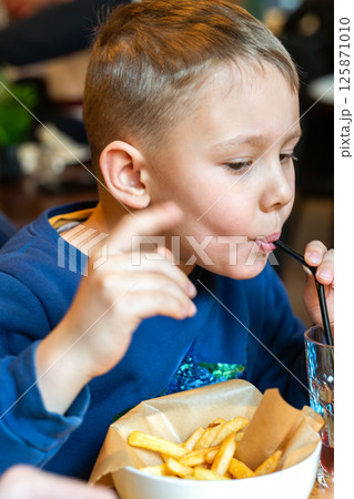 Young boy eating French fries and sipping a drink through a straw at a restaurant table close-up view, concept of casual dining 125871010