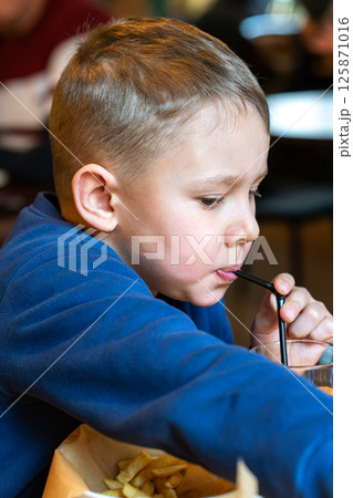 Young boy eating French fries and sipping a drink through a straw at a restaurant table close-up view, concept of casual dining 125871016