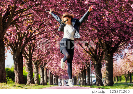 Dynamic scenic young adult pretty happy woman enjoy walking spring city street blooming pink cherry trees alley background in Magdeburg city. Female person joyful carefree portrait smiling outdoors 125871260