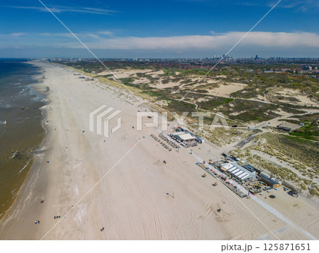 Aerial view of a wide sandy beach with beach bars and dunes, stretching along the coast with a distant city skyline visible under a bright blue sky. 125871651