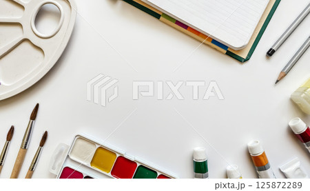 Creative workspace with watercolor paints, paintbrushes, color tubes, sketchbooks, and pencils arranged around a white background 125872289
