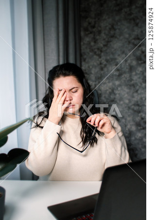 Symptoms of overworking. Young tired female freelancer in eyeglasses sitting in front of computer rubbing eyes feeling eye strain during work 125874924