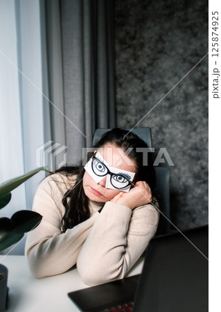 Overworking at office concept. No time to sleep. Female office worker with painted eyes on stickers imitating working in front of computer while sleeping sitting at desk 125874925