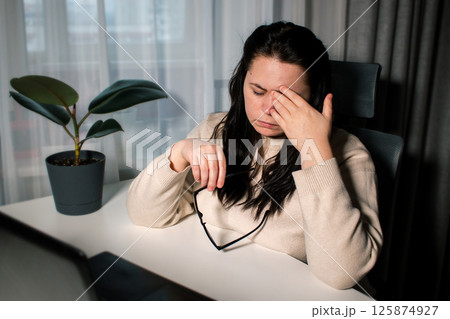 Symptoms of overworking. Young tired female freelancer in eyeglasses sitting in front of computer rubbing eyes feeling eye strain during work Symptoms of overworking. Young tired female freelancer in eyeglasses sitting in front of computer rubbing eyes feeling eye strain during work 125874927