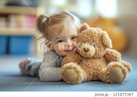 Child lying on a carpet cuddling a soft toy bear in a cozy, blurred background setting. 125875367