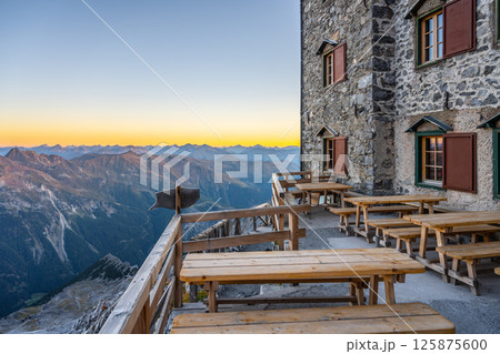 Visitors enjoy breathtaking mountain views from the terrace of Julius Payer House. The surrounding Ortler Alps showcase stunning peaks at sunset, creating a picturesque backdrop. 125875600
