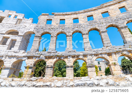 This remarkable Roman amphitheatre in Pula showcases its grand arches against a bright blue sky. Visitors can imagine the vibrant history of events that once took place here. 125875605