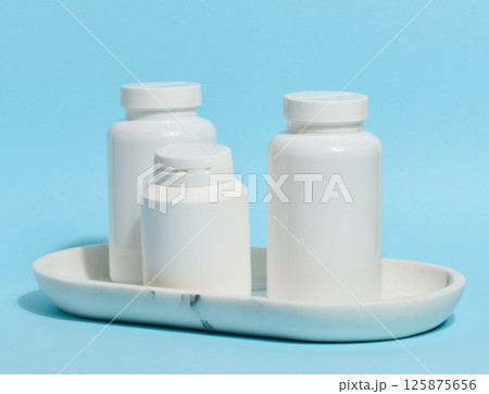 Three white plastic pill bottles of varying sizes stand on a white marble tray against a light blue background 125875656
