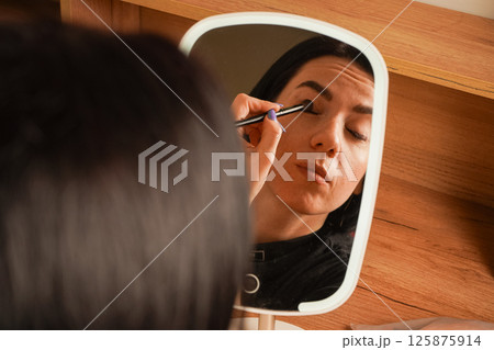 A young woman applies makeup, carefully lining her eyes in front of a mirror. Her black hair frames her face as she focuses on her reflection. Behind her is a cozy wooden shelf with decor. 125875914