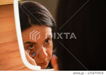 Behind her is a cozy wooden shelf with decor. A young woman applies makeup, carefully lining her eyes in front of a mirror. Her black hair frames her face as she focuses on her reflection. Behind her is a cozy wooden shelf with decor. A young woman applies makeup, carefully lining her eyes in front of a mirror. Her black hair frames her face as she focuses on her reflection. 125875919