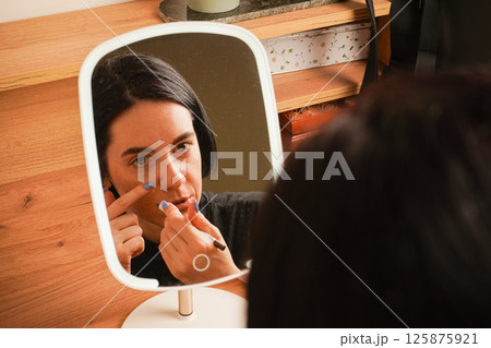 A young girl sits near a small mirror. Attributes for applying makeup next to the girl. In the mirror, a reflection of a girl with short black hair. 125875921