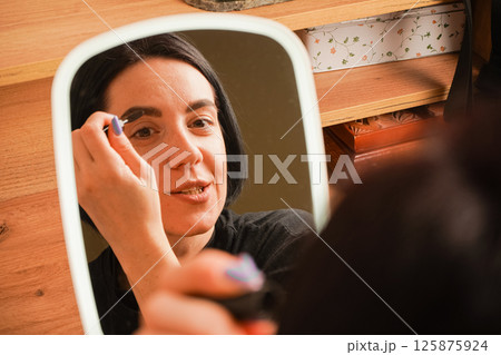 Applying mascara to her brows, a woman admires her reflection in a hand-held mirror. Her short black hair adds elegance to her look. The room is accented by warm wooden tones. 125875924