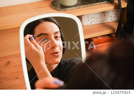 Her short black hair adds elegance to her look. The room is accented by warm wooden tones. Applying mascara to her brows, a woman admires her reflection in a hand-held mirror. Her short black hair adds elegance to her look. The room is accented by warm wooden tones. Applying mascara to her brows, a woman admires her reflection in a hand-held mirror. 125875925