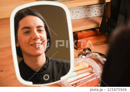 A young girl sits near a small mirror. Attributes for applying makeup next to the girl. In the mirror, a reflection of a girl with short black hair. A young girl sits near a small mirror. Attributes for applying makeup next to the girl. In the mirror, a reflection of a girl with short black hair. 125875926