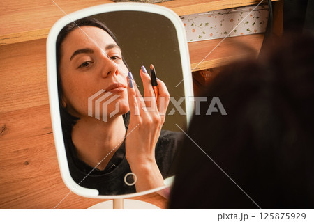 Her relaxed expression reflects self-care. Behind her, a wooden shelf and small decorative items create a cozy atmosphere. A close-up of a woman applying makeup shows her lining her lips. 125875929