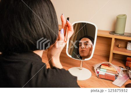 In the mirror, a reflection of a girl with short black hair. A young girl sits near a small mirror. Attributes for applying makeup next to the girl. In the mirror, a reflection of a girl with short black hair. A young girl sits near a small mirror. Attributes for applying makeup next to the girl. 125875932