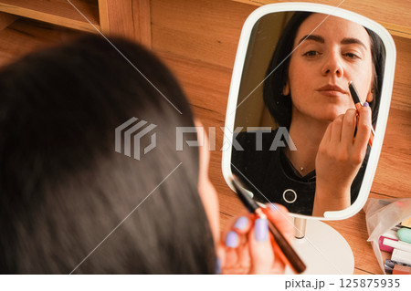 Her short black hair adds elegance to her look. The room is accented by warm wooden tones. Applying mascara to her brows, a woman admires her reflection in a hand-held mirror. 125875935