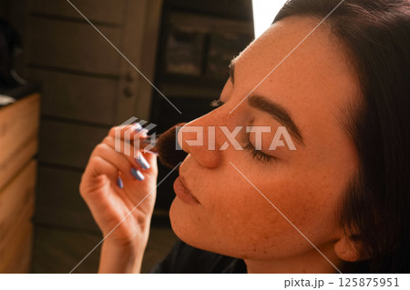 A young woman gently applies powder to her face using a makeup brush. Her eyes are closed, highlighting her focus on self-care. Her black bob haircut and natural complexion stand out. 125875951