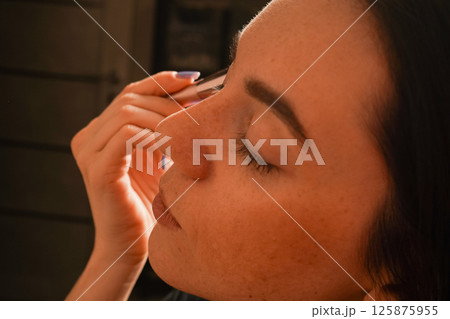 Her eyes are closed, highlighting her focus on self-care. Her black bob haircut and natural complexion stand out. A young woman gently applies powder to her face using a makeup brush. 125875955