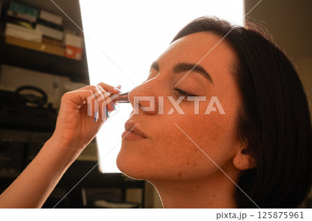 Her eyes are closed, highlighting her focus on self-care. Her black bob haircut and natural complexion stand out. A young woman gently applies powder to her face using a makeup brush. Her eyes are closed, highlighting her focus on self-care. Her black bob haircut and natural complexion stand out. A young woman gently applies powder to her face using a makeup brush. 125875961