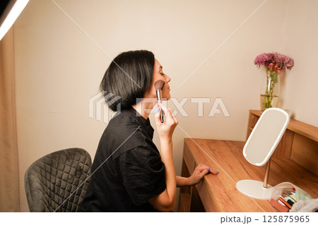 A woman carefully applies powder to her face with a large makeup brush. Her natural beauty and black hair shine in the warm light. The woman sits at a makeup table with a small mirror in front of her. A woman carefully applies powder to her face with a large makeup brush. Her natural beauty and black hair shine in the warm light. The woman sits at a makeup table with a small mirror in front of her. 125875965
