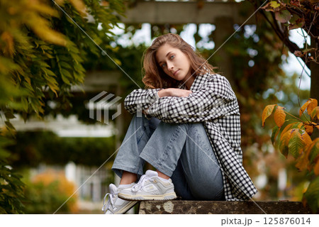 Stylish young woman in casual autumn outfit sitting on a stone wall in a green park with orange leaves, wearing a black and white plaid shirt, jeans, and sneakers – outdoor fashion portrait. Stylish young woman in casual autumn outfit sitting on a stone wall in a green park with orange leaves, wearing a black and white plaid shirt, jeans, and sneakers – outdoor fashion portrait. 125876014