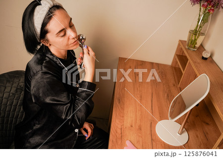 A girl in black pajamas sits at a table in front of a mirror. Her expression reflects self-care. A woman uses a delicate metal roller to massage her jawline for a soothing skincare treatment. A girl in black pajamas sits at a table in front of a mirror. Her expression reflects self-care. A woman uses a delicate metal roller to massage her jawline for a soothing skincare treatment. 125876041