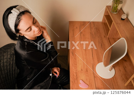 Her expression reflects self-care. A woman uses a delicate metal roller to massage her jawline for a soothing skincare treatment. A girl in black pajamas sits at a table in front of a mirror. 125876044
