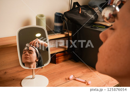 Girl with a headband holding her hair. Young woman doing anti-aging facial massage with a metal roller. Reflection of brunette during massage in table mirror. 125876054