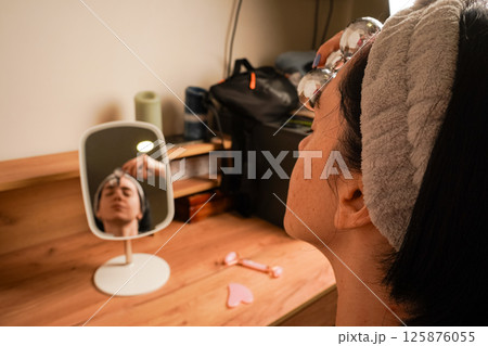 Young woman doing anti-aging facial massage with a metal roller. Reflection of brunette during massage in table mirror. Girl with a headband holding her hair. Young woman doing anti-aging facial massage with a metal roller. Reflection of brunette during massage in table mirror. Girl with a headband holding her hair. 125876055