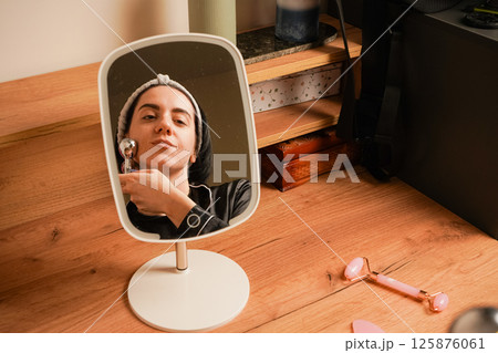 Young woman doing anti-aging facial massage with a metal roller. Reflection of brunette during massage in table mirror. Girl with a headband holding her hair. Young woman doing anti-aging facial massage with a metal roller. Reflection of brunette during massage in table mirror. Girl with a headband holding her hair. 125876061