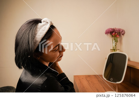 Reflection of brunette during massage in table mirror. Girl with a headband holding her hair. Young woman doing anti-aging facial massage with a metal roller. Reflection of brunette during massage in table mirror. Girl with a headband holding her hair. Young woman doing anti-aging facial massage with a metal roller. 125876064
