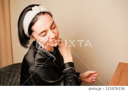 Her black silk pajamas add elegance to the scene. The soft lighting enhances the calming atmosphere. Performing a gua sha facial massage, a woman focuses on her face and neck. 125876156