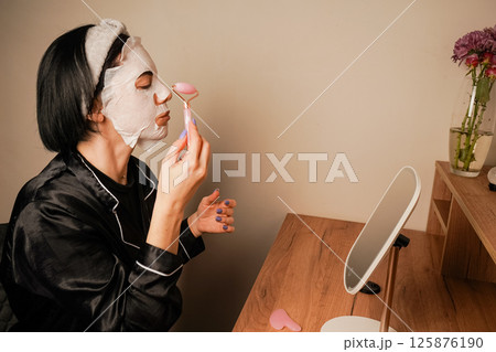 A small mirror adds charm to the scene. A young woman is using a hydrating mask at her dressing table. She is wearing elegant black pajamas and a headband. 125876190