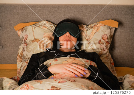 Her black pajamas and soft floral bedding create a serene atmosphere. Her relaxed posture highlights a restful sleep. Peacefully lying in bed, a woman enjoys her rest with a silk eye mask. 125876270