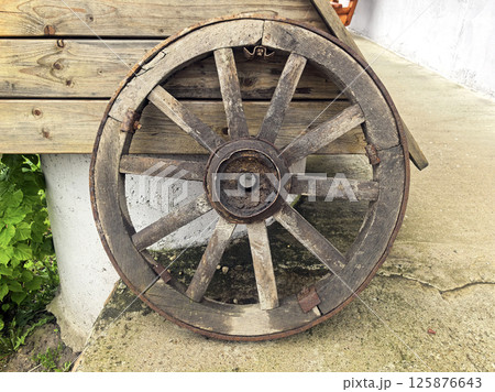 An old wooden cart wheel stands on a concrete path against a background of planks. Transportation used in villages, ranches, and farms. Vintage and antique An old wooden cart wheel stands on a concrete path against a background of planks. Transportation used in villages, ranches, and farms. Vintage and antique 125876643