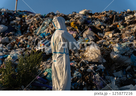 Individual wearing a protective suit stands thoughtfully in front of a vast mountain of waste in a sunny urban setting Individual wearing a protective suit stands thoughtfully in front of a vast mountain of waste in a sunny urban setting 125877216