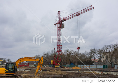 A tall red crane is actively lifting materials over a construction site while heavy machinery is positioned nearby, under a cloudy sky A tall red crane is actively lifting materials over a construction site while heavy machinery is positioned nearby, under a cloudy sky 125877272