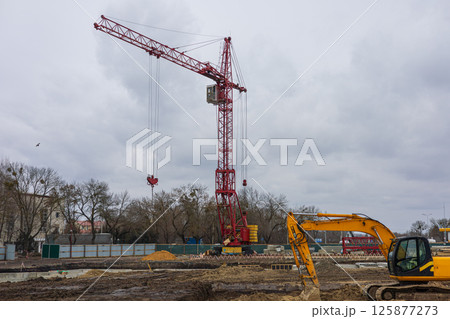 Heavy machinery operates at a construction site featuring a prominent red crane against a gray, cloudy sky, with trees in the background 125877273