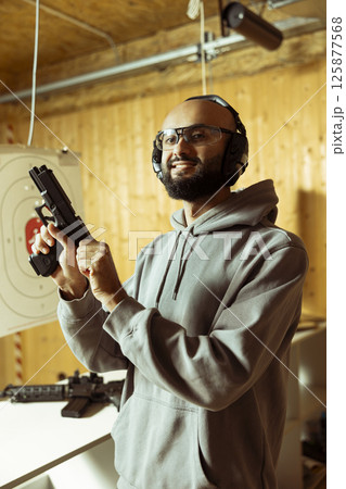 Portrait of happy Arab man in shooting range cocking handgun, setting mechanism up for firing. Cheerful Middle Eastern person pulling back pistol hammer during training session, preparing to shoot Portrait of happy Arab man in shooting range cocking handgun, setting mechanism up for firing. Cheerful Middle Eastern person pulling back pistol hammer during training session, preparing to shoot 125877568