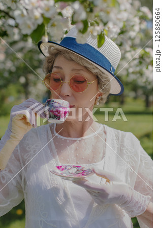 Happy natural adult mature woman of 60 years old in white canotier hat and red glasses, retro dress, gloves drinks tea in blossoming white apple orchard on summer day Happy natural adult mature woman of 60 years old in white canotier hat and red glasses, retro dress, gloves drinks tea in blossoming white apple orchard on summer day 125880664