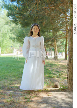 A young lady in a white outfit is preparing for her First Communion. The girl looks solemn against a background of emerald coniferous branches. 125881705