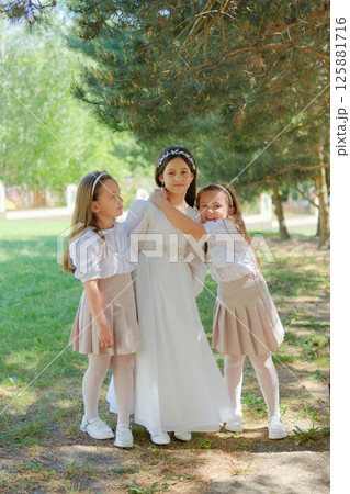 Three girls in festive attire stand against a backdrop of emerald trees. The central girl in an ivory dress radiates calm, with two younger twins nearby. Three girls in festive attire stand against a backdrop of emerald trees. The central girl in an ivory dress radiates calm, with two younger twins nearby. 125881716