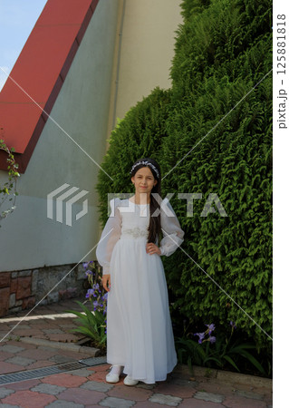 A young girl in a long dress poses in front of the stone walls of a church. The outfit emphasizes the solemn mood and purity of intentions. 125881818
