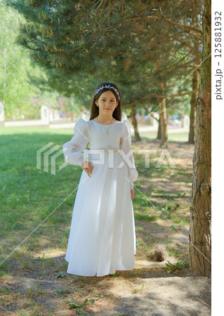A girl in a milky holiday outfit stands under the pine trees. The little lady looks like a fairy-tale heroine among the green silence.  125881932