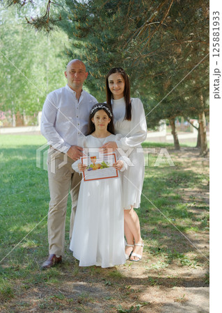 A little girl on the eve of her First Communion looks solemn next to her father and mother. The father gently leans toward the girl, and the mother gently hugs her shoulders.  125881933