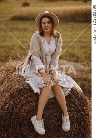 Young woman in beige dress sitting on haystack in village field Young woman in beige dress sitting on haystack in village field 125882299